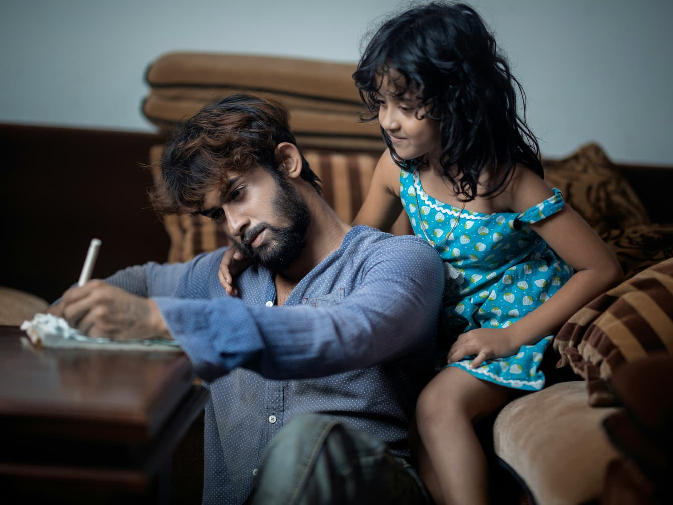 A father writes at a table while his young daughter leans on his shoulder and watches.