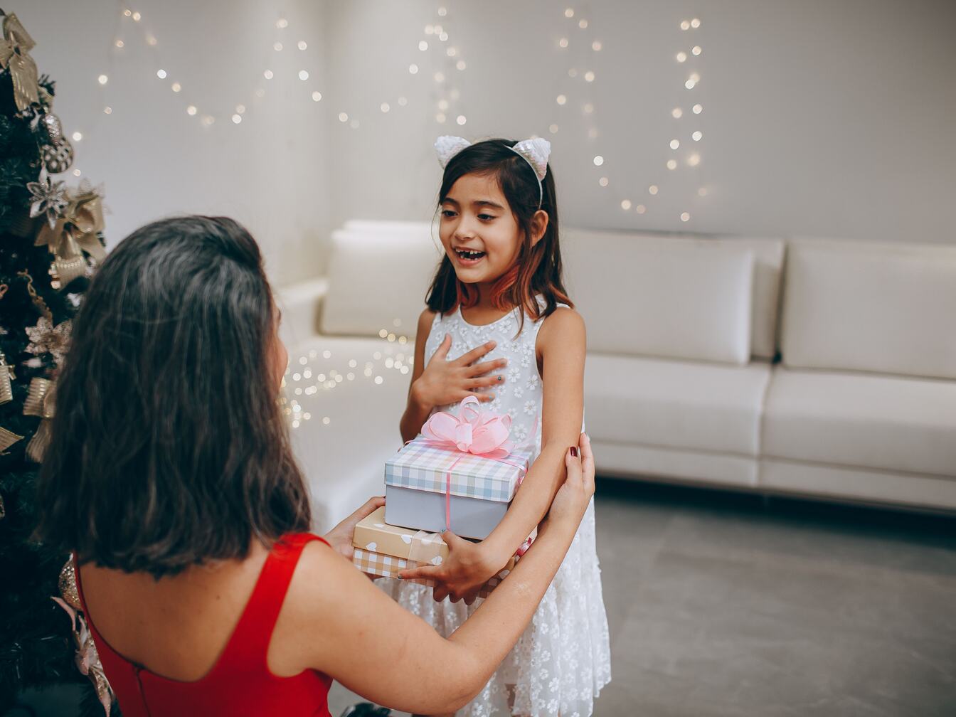 A delighted girl holds a stack of wrapped presents, her hand on her heart, as her mother kneels to give them.