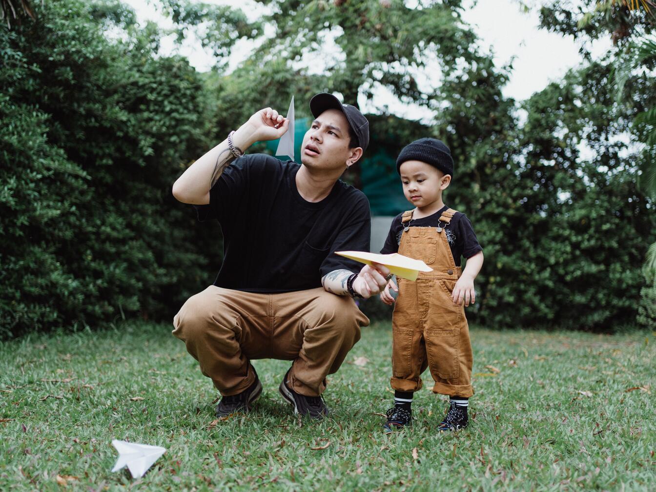 A father crouches on a lawn beside his toddler in brown overalls, both about to launch paper airplanes.