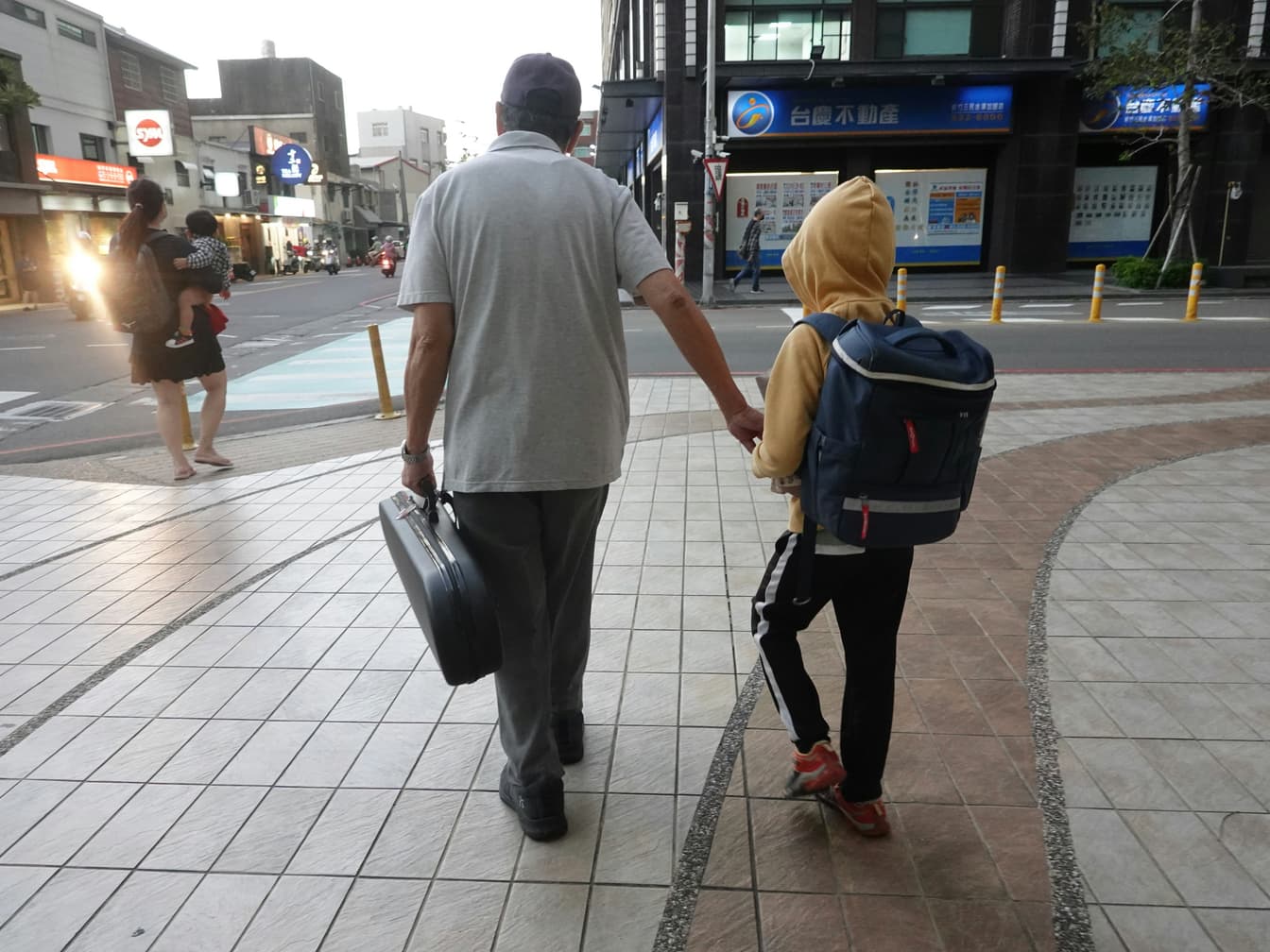 An older man carrying a briefcase walks hand in hand with a child wearing a school backpack along a city sidewalk.
