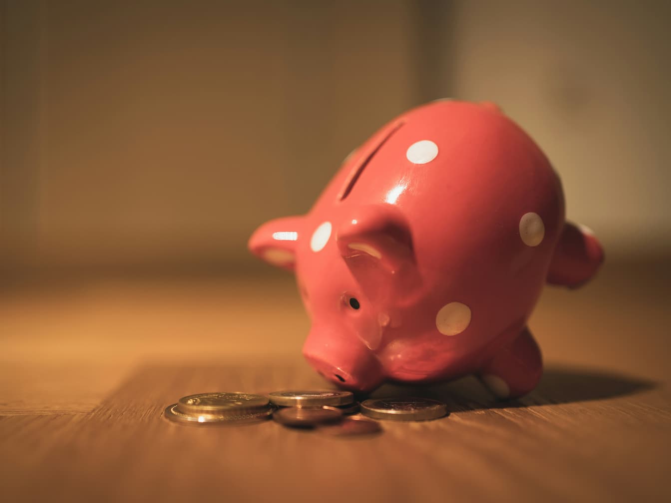 A pink polka-dot piggy bank on a wooden table, with a few coins scattered in front of it.
