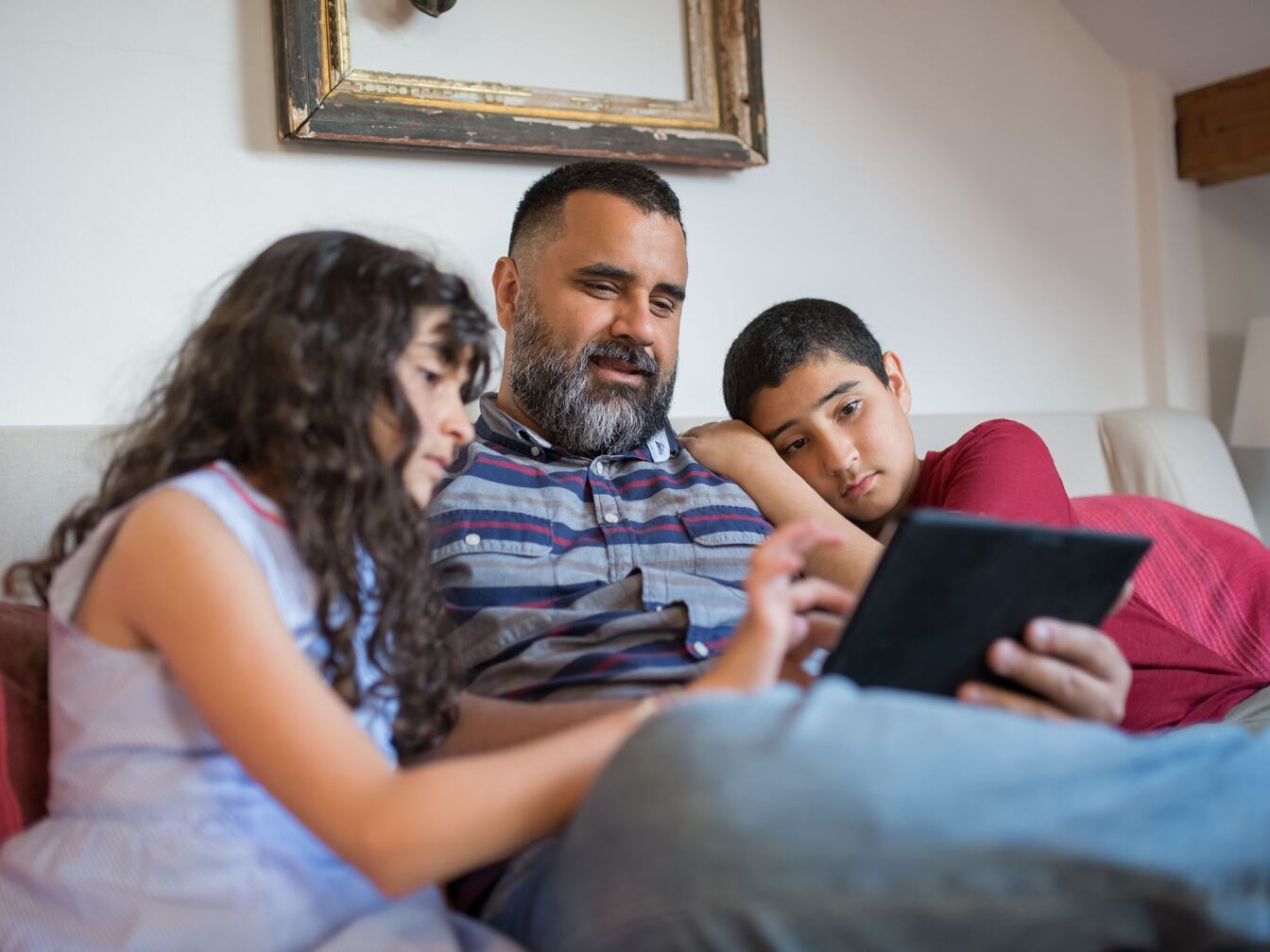 A father sits on a sofa with his son and daughter as she points at something on a tablet.