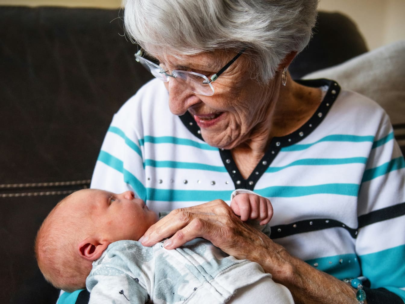 A great-grandmother in a striped sweater smiles down at the newborn she is holding on her lap.