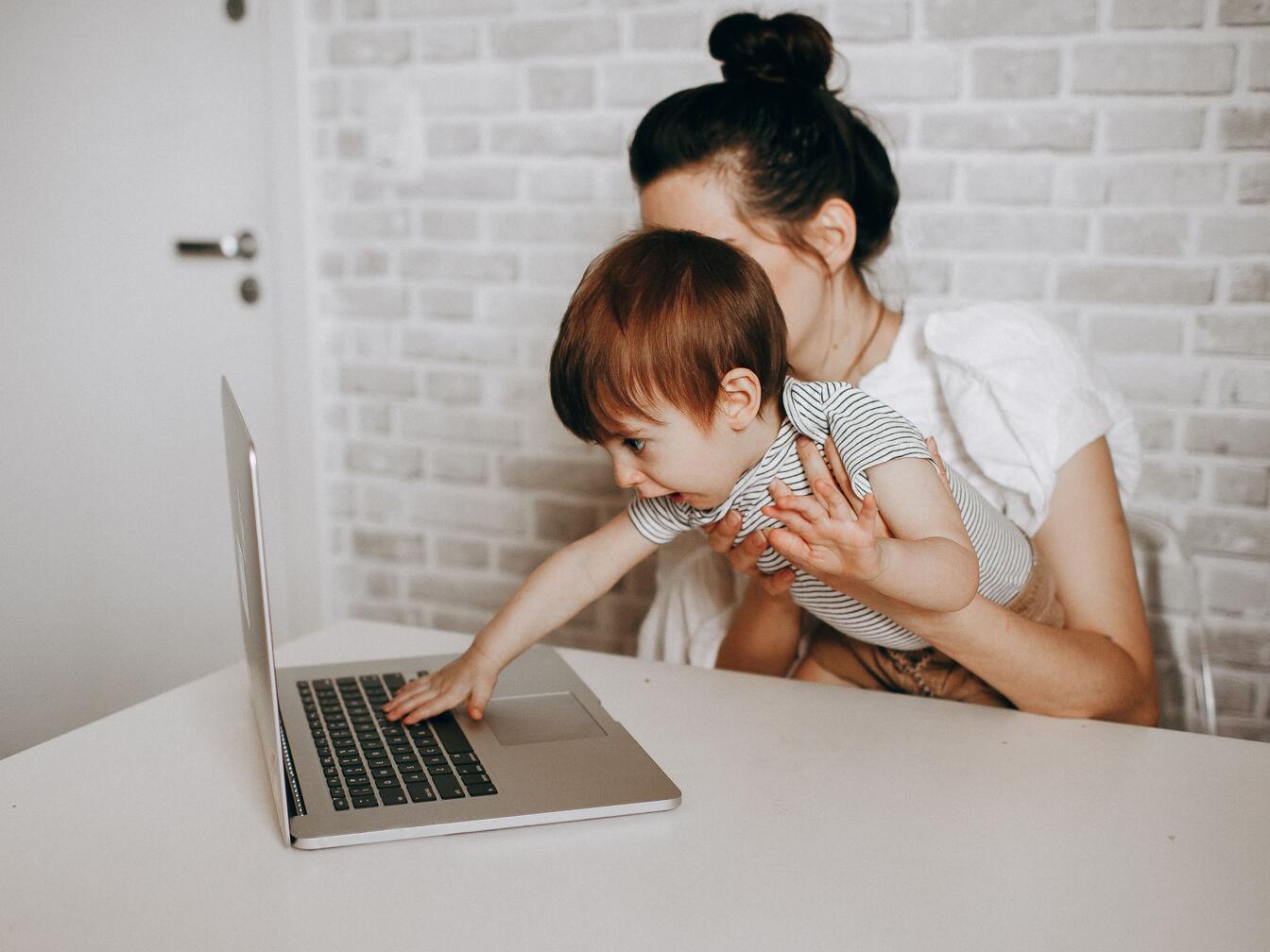 A baby on his mother’s lap leans forward to tap the keys of a laptop on the table.