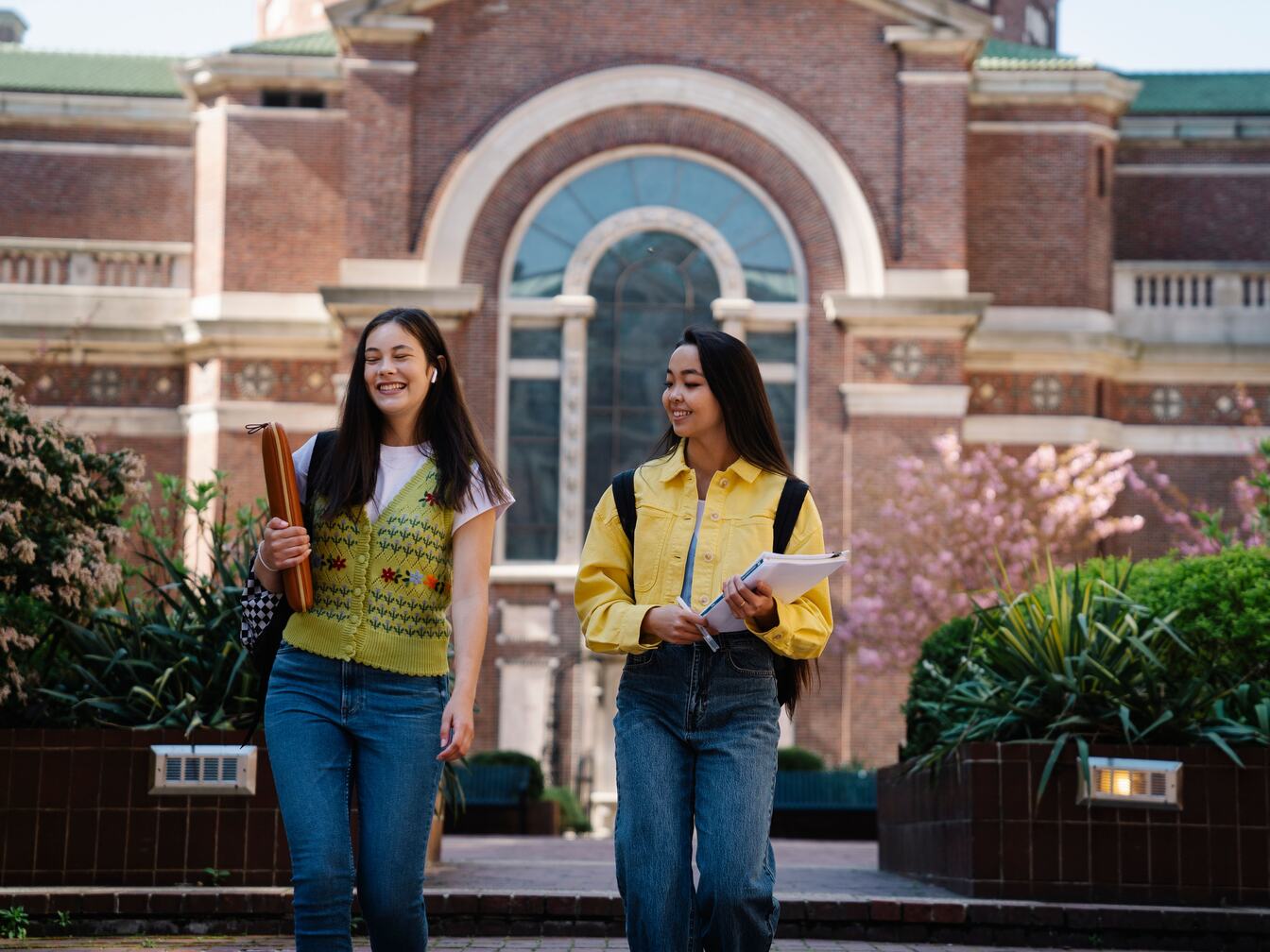 Two smiling college students with backpacks and notebooks walk past a brick campus building.