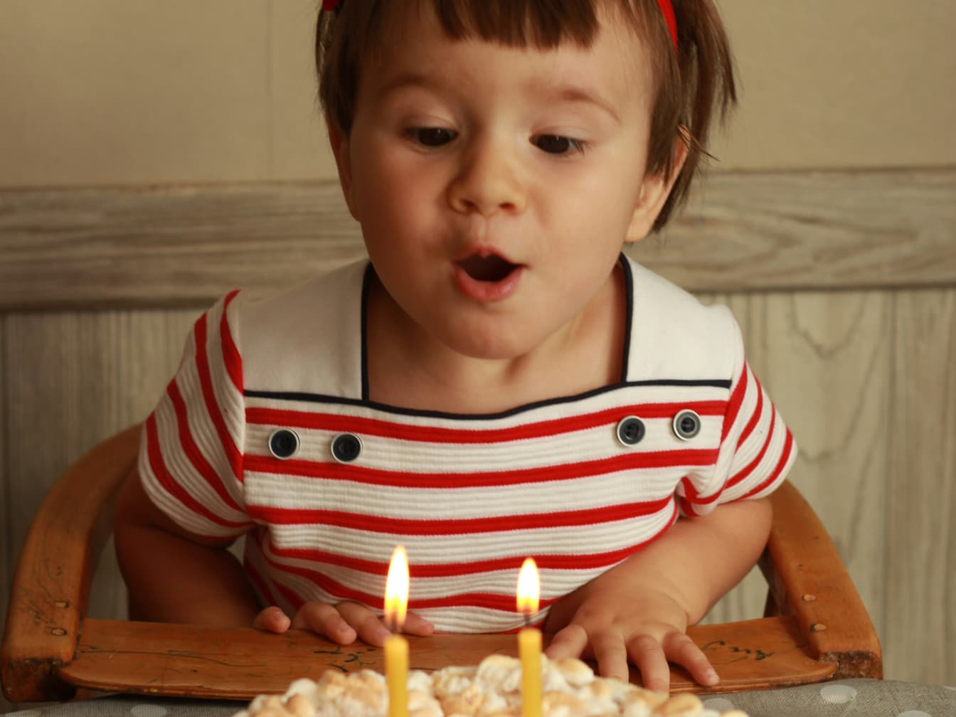 A toddler in a striped top leans forward to blow out two candles on a birthday cake.