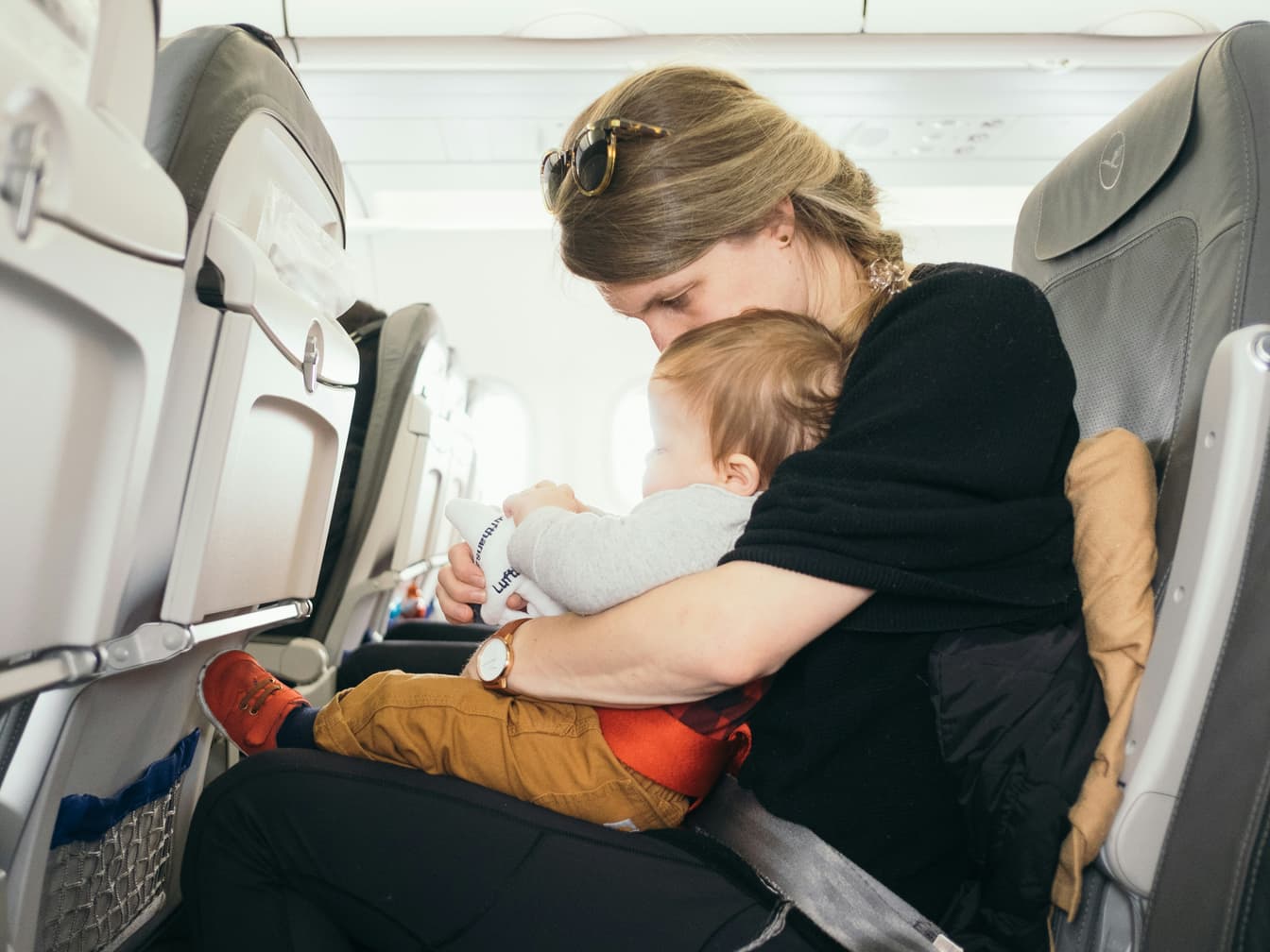 A mother in an airplane seat holds her baby close, her cheek resting on the baby’s head.