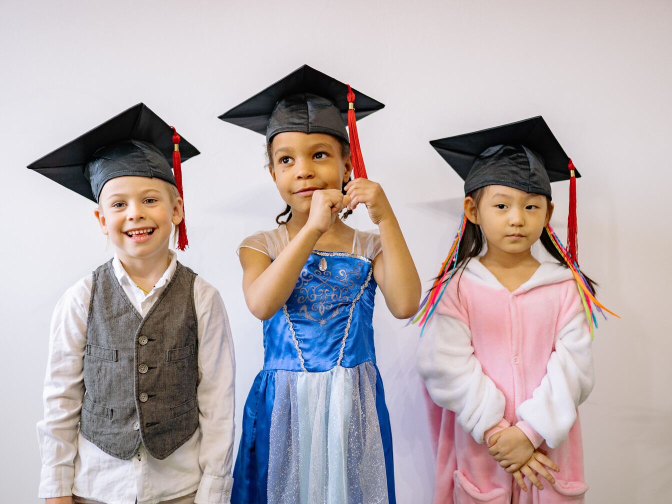 Three small children in black graduation caps stand in a row, one twirling her red tassel.
