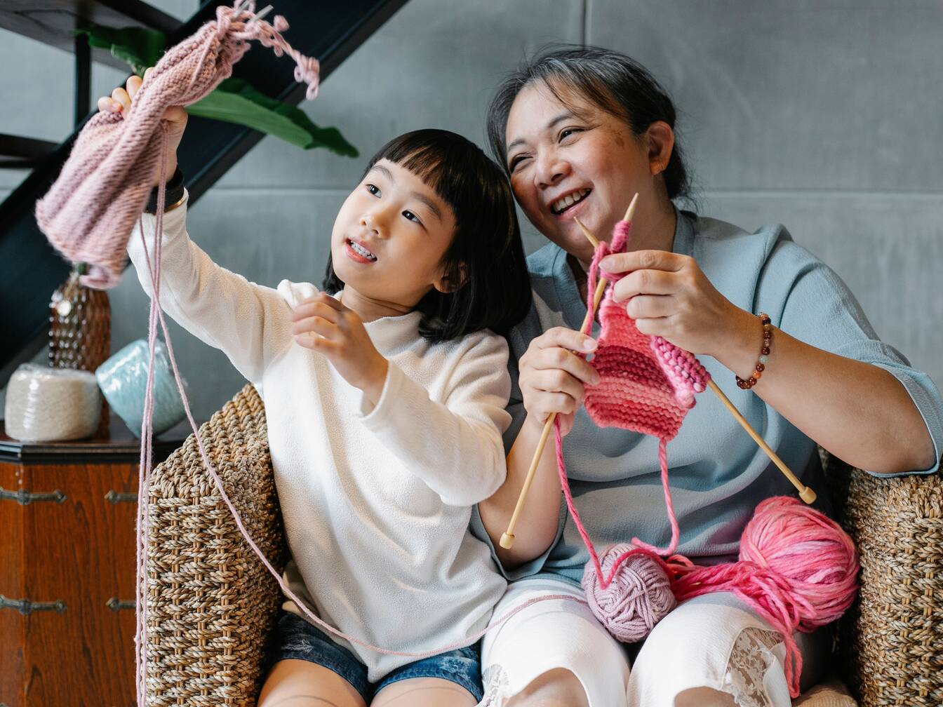 A young girl proudly holds up her knitting while her grandmother laughs beside her, needles and yarn in hand.