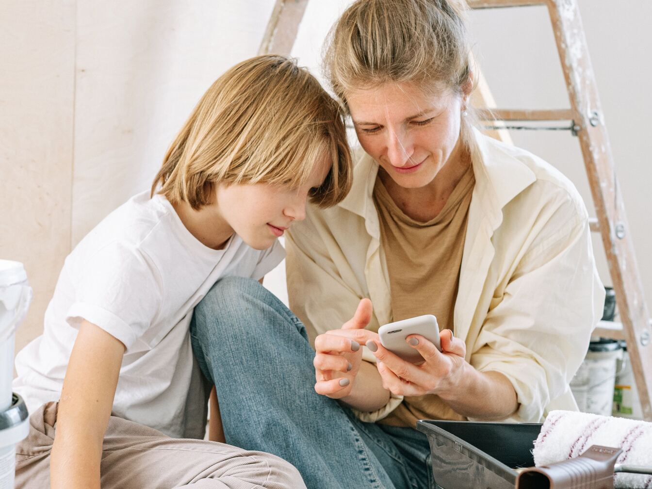 A mother shows her young teenage son something on her phone as they sit close together on the floor.