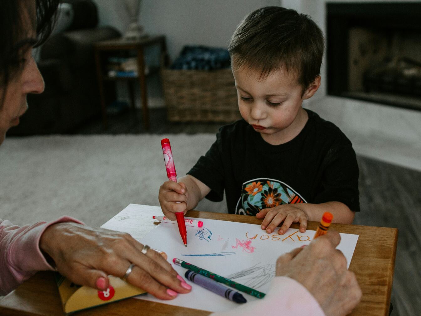 A little boy concentrates as he writes with a red marker, while an adult beside him adds to the page.
