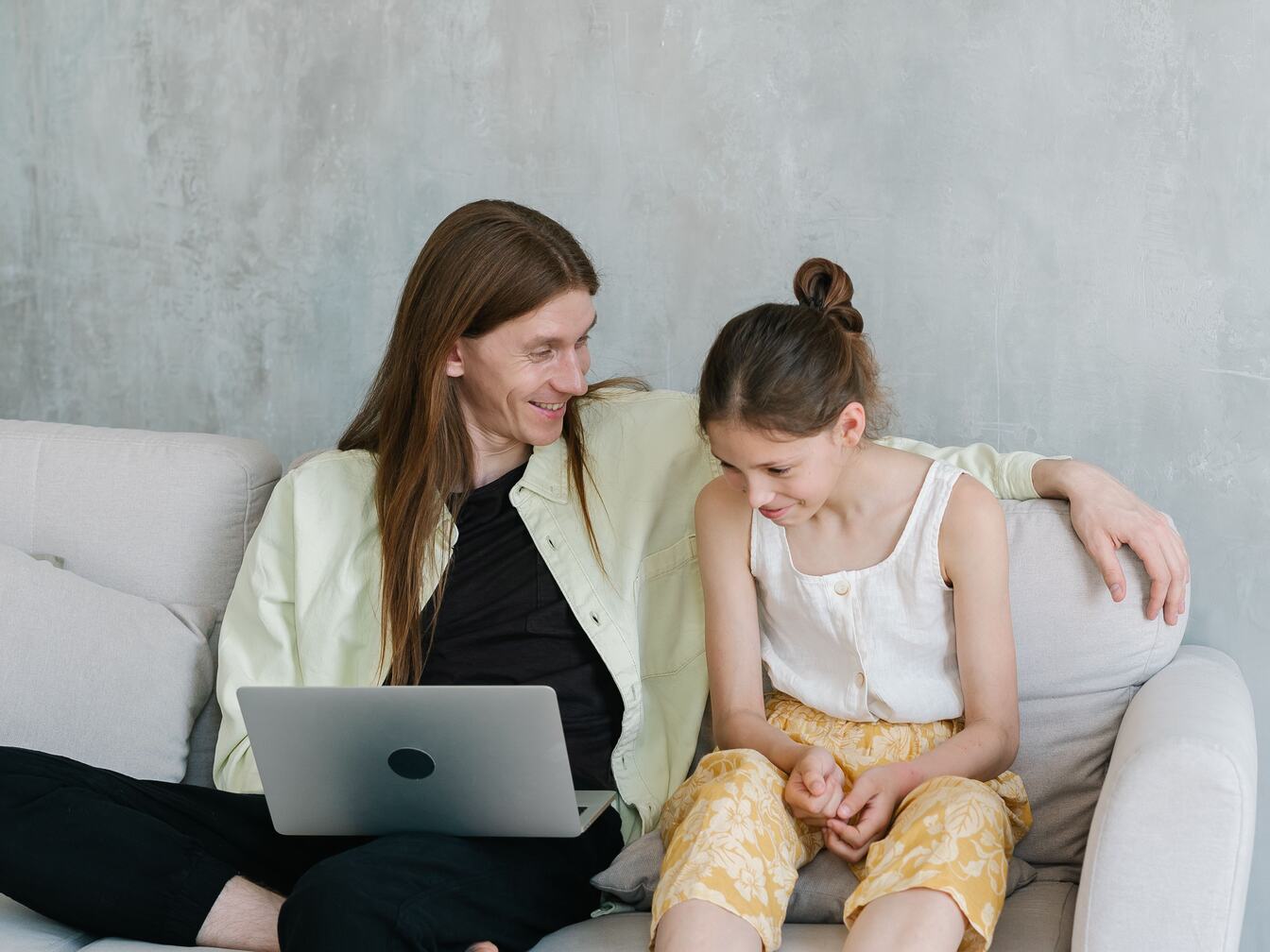 A father sits on a sofa with a laptop, his arm around his smiling daughter as they look at the screen together.