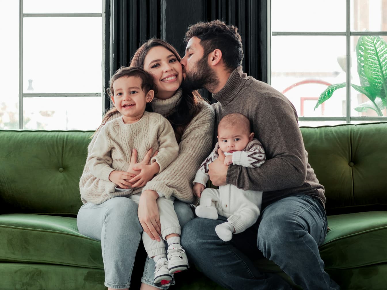 Two parents sit close on a green sofa, one holding a smiling toddler and the other a baby, as the father kisses the mother’s cheek.