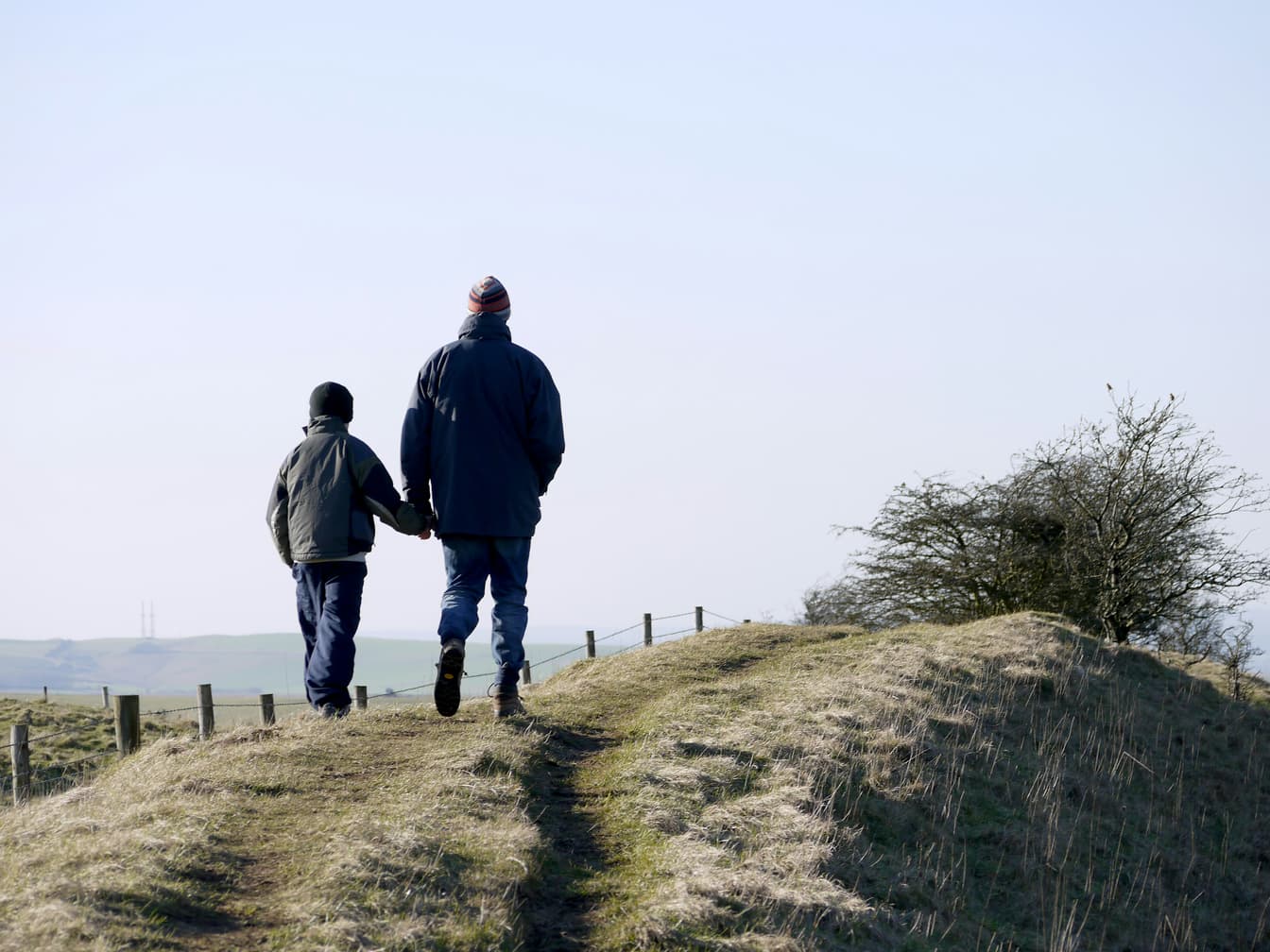 A man and a boy walk side by side up a grassy hill path under a clear sky.